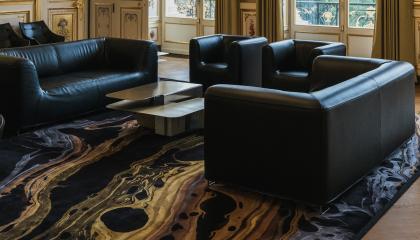 Huoneessa lattialla tumma matto, jossa on vaaleita kuvioita. Huoneessa keskellä on tumma sohvaryhmä ja nojatuolit.