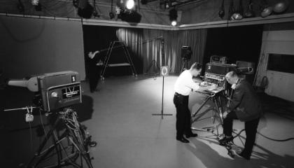 Kaksi henkilöä seisoo televisiolaitteen ääressä studiossa. Etualalla on videokamera.