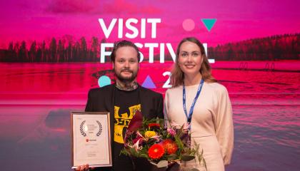 Palkinnon voittaja poseeraa kukkien ja taulun kanssa kaupungin edustajan kanssa. 