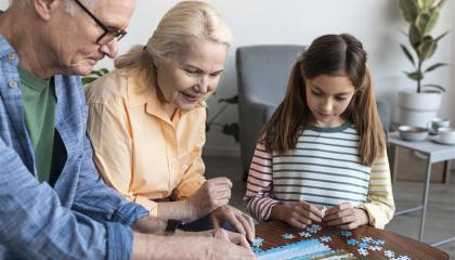 Isoäiti ja isoiso istuvat sohvapöydän ääressä kokoamassa palapeliä tummahiuksisen lapsen kanssa.