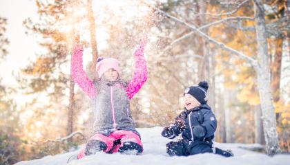 Kuvassa kaksi lasta istuu lumihangessa ja heittää lunta.