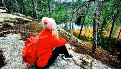 Punatakkinen tyttö reppu selässään istuu kalliolla ja katselee maisemaa, jossa näkyy metsää ja pieni järvi.