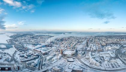 Ilmakuvassa levittäytyy luminen Tampere. Keskellä Nokia Arena ja risteilevät tiet, taustalla järvet ja sininen taivas.