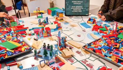 Isolla pöydällä on kasa värikkäitä lego-palikoita, joista ihmisiä on rakentamassa karttapohjalle toiveidensa asuinaluetta.