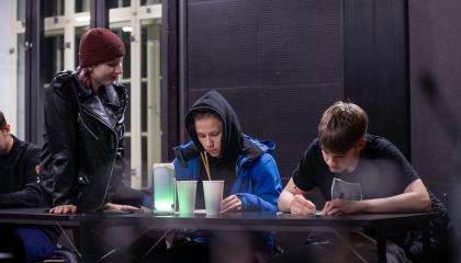 Kaksi nuorta istuu pöydän ääressä kirjoittamassa ja yksi nuori istuu pöydällä ja hymyilee