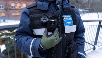 Matkalippujen tarkastaja seisoo ja esittelee varusteiden haalarikameraa.