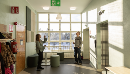 Kaksi henkilöä katsoo ylöspäin kohti kattovalaisimia koulun käytävän päässä ikkunan edessä.