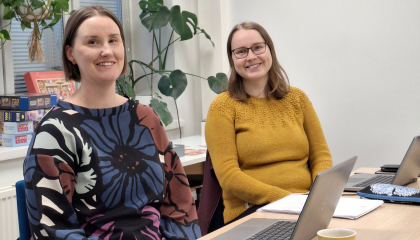 Kuvassa kaksi naista istumassa pöydän ääressä. Molemmat hymyilevät.