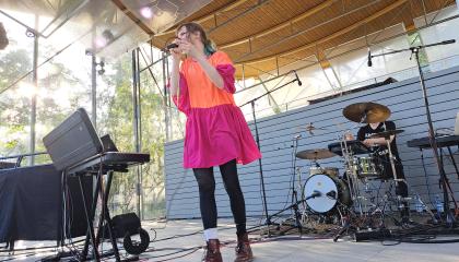 Pinkki-oranssiin mekkoon pukeutunut laulaja esiintyy ja mustiin pukeutunut rumpali soittaa taustalla.