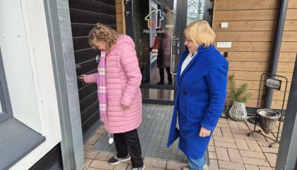A woman with an intellectual disability, dressed in a pink padded jacket, is unlocking the front door of her home. Her mother, dressed in blue, is standing behind her.
