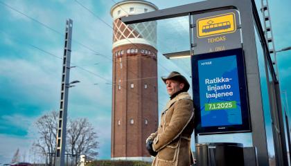 Cowboy-hattuinen mies nojaa pysäkkikatokseen, jossa näkyy mainos: Ratikkalinjasto laajenee 7.1.2025.