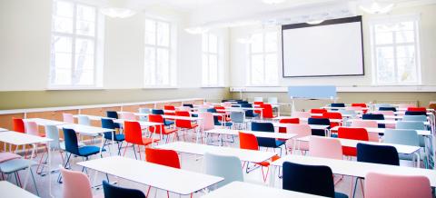 The bright lecture hall has rows of white tables and colourful chairs with velvet cushions. The back wall of the room is covered with a screen.