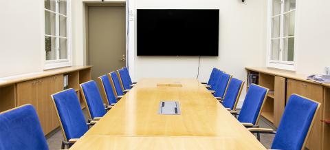 The elongated space features a light wood table that fills the entire space, surrounded by blue chairs with backrests. On the back wall is a large television screen and a door.