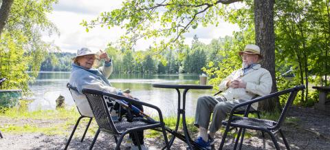 Ikääntynyt pariskunta aurinkoisena kesäpäivänä istumassa Riihiniemen uimarannalla pienen pöytäryhmän ääressä.