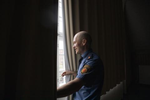 Mika katsoo ikkunasta verhojen välistä ulos. Hänellä on pelastuslaitoksen paita päällä.