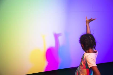 Child holding one hand up, in front of child is colorful wall.