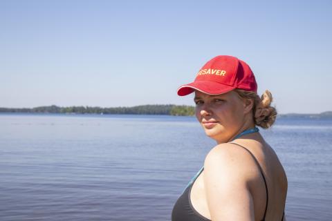 Uimavalvoja punaisessa lippalakissa katsoo kameraa. Taustalla näkymä järvelle.