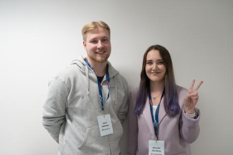 Nuori poika vasemmalla, lyhyet vaaleat hiukset. Nuori violettitukkainen nainen oikealla.