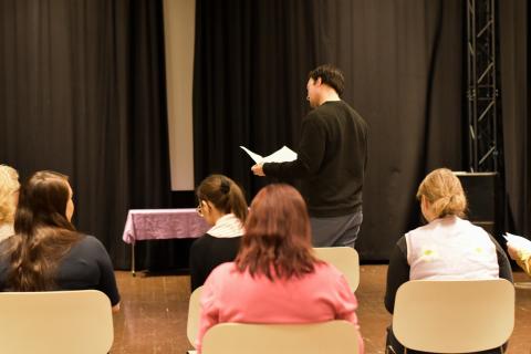 Henkilö lukee taustalla käsikirjoitusta. Edessä rivissä opiskelijoita.