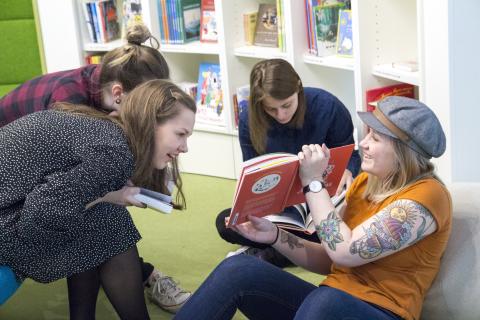 Neljä nuorta kirjastossa lukemassa. Yksi heistä näyttää muille avointa kirjaa.
