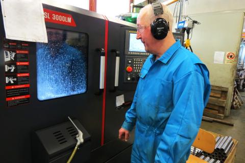 Mies seisoo ison koneen edessä. Hän katsoo sen näyttöä. Hänellä on sininen haalari päällä, suojalasit ja kuulosuojamet.