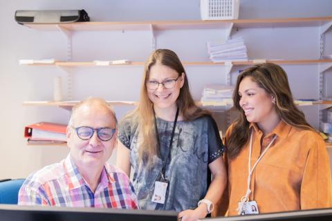 Rakennusvalvojat katsovat samaa tietokonenäyttöä hymyillen. Takana työhuoneen seinähylly.