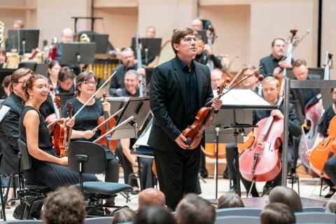 The Tampere Philharmonic Orchestra's musicians sitting with their instruments in the concert hall, Johannes Põlda standing with his violin and facing the audience.