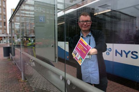 Mikko Ala-Kapee at a bus stop holding a Let's make Tampere equal together -poster, with a bus in the background.