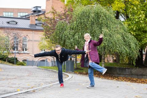 Kaksi henkilöä seisoo yhdellä jalalla tasapainoillen puiden ja punatiilisen rakennuksen edessä..