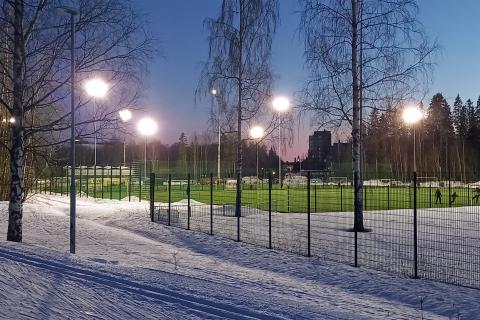 Kaupin hiihtoladun hämärtyvässä illassa. Taustalla jalkapalloilijoita tekonurmikentällä, jossa valot loistavat.