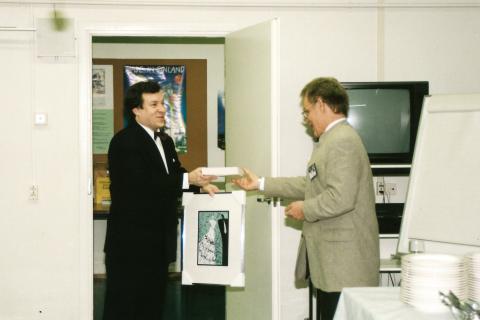 Henkilö ojentaa toiselle henkilölle lahjan. Lahjansaajalla on kädessään taulu.