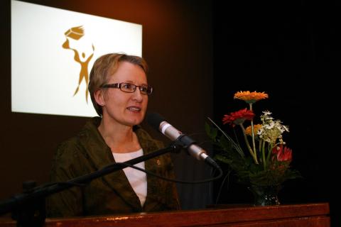 Henkilö seisoo puhujanpöntössä. Edessä on mikrofoni ja vieressä kukkia.