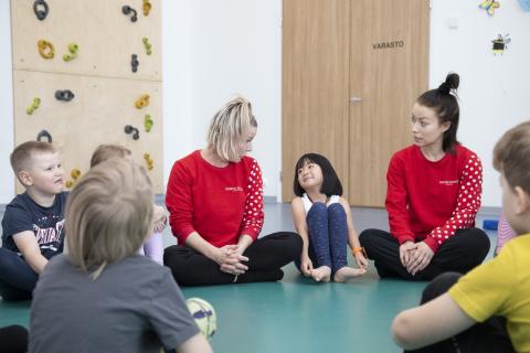 Lapsiryhmä ja kaksi sirkusopettajaa istuu liikuntasalin lattialla juttelemassa. 