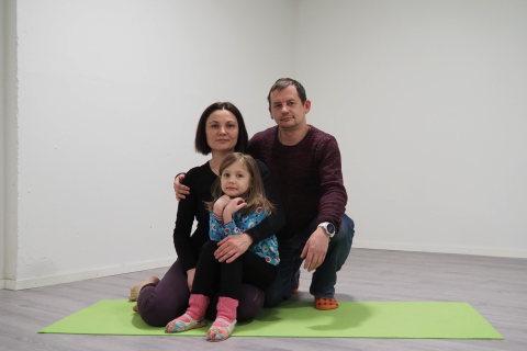 Kolmihenkinen perhe istuu vihreän jumppamaton päällä. Lapsi istuu äidin sylissä.