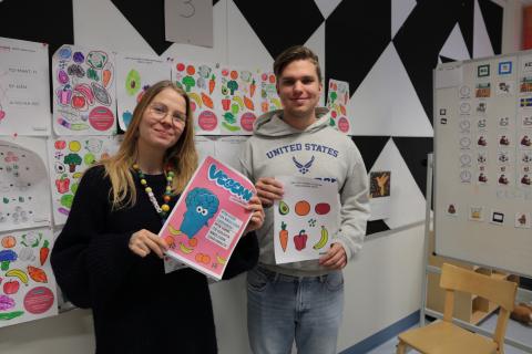 Two people holding Vegemi material and smiling. Behind them is a wall full of colouring pictures of fruits and vegetables..