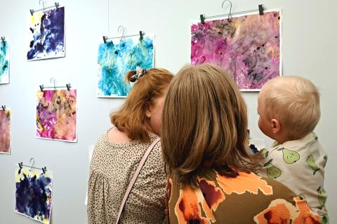 Seinällä abstrakteja vauvojen ja taaperoiden tekemiä taideteoksia. Kaksi aikuista ja toisella vauva sylissään ihastelemassa teoksia. 