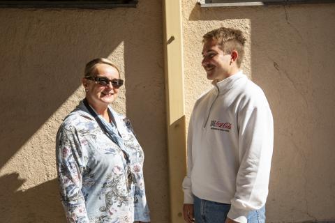 A woman and a man stand side by side on the wall of a light-coloured building. She looks at the camera, he looks at her.