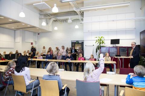 Suuressa salissa istuu paljon lapsia pöydän ääressä. He katselevat salin edessä seisovaa pormestaria, joka hymyilee. Yksi lapsista viittaa kädellään kysyäkseen jotakin.