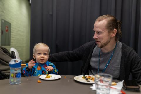 Isä ja pikkulapsi istuvat ruokalassa pöydän ääressä. Lapsi syö sormin ruokaansa. Isän käsi on lapsen olkapäällä.