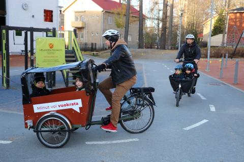 Kaksi henkilöä pyöräilee kahdella laatikkopyörällä päiväkodin pihassa.