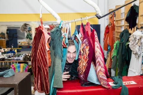 Hymyilevä henkilö kurkistaa tangolla roikkuvien värikkäiden vaatteiden välistä. Vieressä oleville puolapuillekin on ripustettu vaatteita.