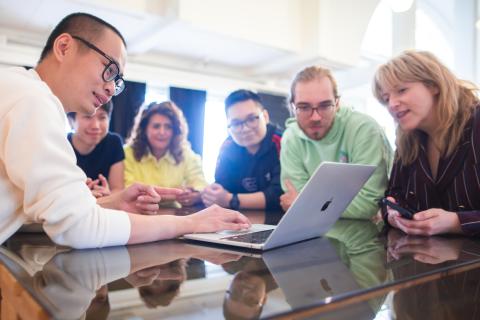 Kuusi henkilöä iloisesti katsomassa tietokoneen näyttöä valoisassa toimistotilassa.