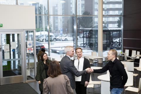 Kaksi henkilöä kättelee toisiaan avarassa ulkoaulassa ja kolme muuta seuraa tilannetta.