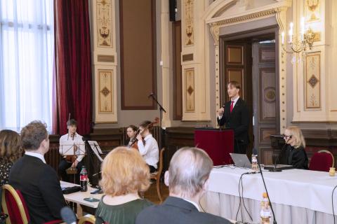 The Council chair gives his speech next to a small orchestra. There are councillors in the foreground photographed from behind.