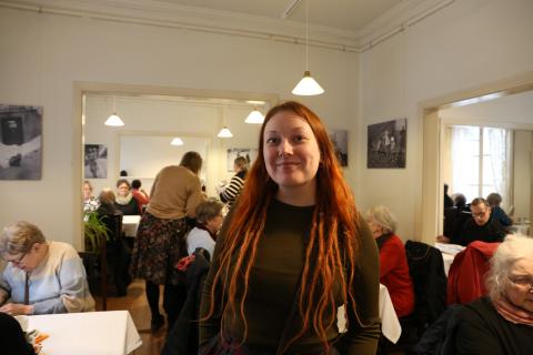 Henkilö edessä hymyilee, taustalla ihmisiä keskustelemassa ja kahvilla.