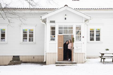 Henneri Play Activity Center in winter. The porch door of the old white wooden house is open. Two friendly-looking adults look on from the door.