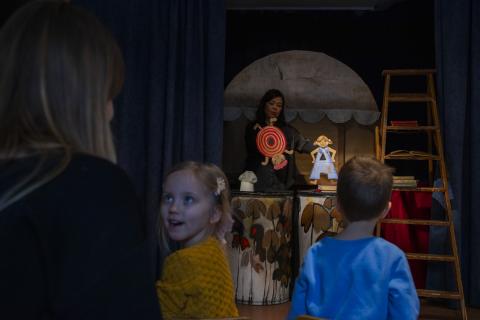 In a dimly lit hall, children watch a paper puppet show. In the foreground, a child watches her mother, enthralled by the theatrical performance.