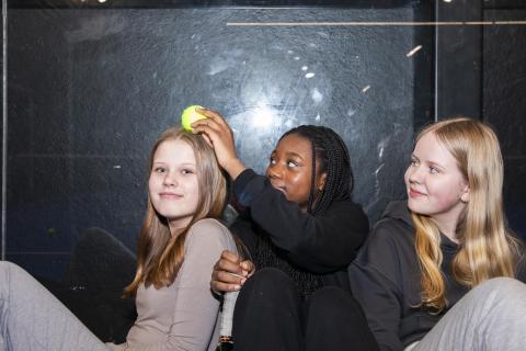 Three youngster sits on the padel floor and one of them holds a tennis ball above another's head.