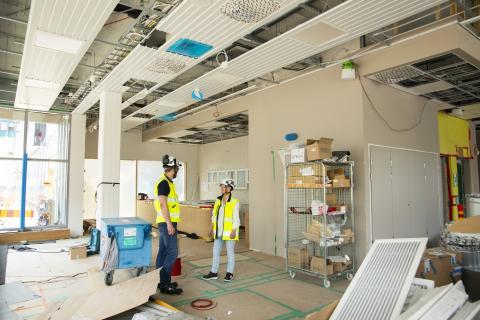 Kaksi työmaaliiveihin ja -kypäriin pukeutunutta henkilöä juttelee tulevissa kahvilan tiloissa. Ympärillä on rakennustyömaan tarvikkeita.