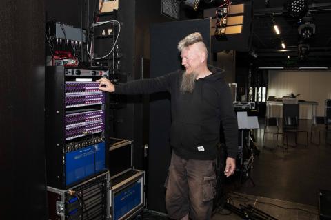 Henkilö seisoo itsensä kokoisen äänimikserin vieressä ja säätää sitä.